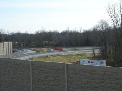 Sound control barrier in the foreground with lower wye tracks leading off to the left (Indiana) and right (Joliet). Former EJ&E track runs from left to right just behind orange fencing.